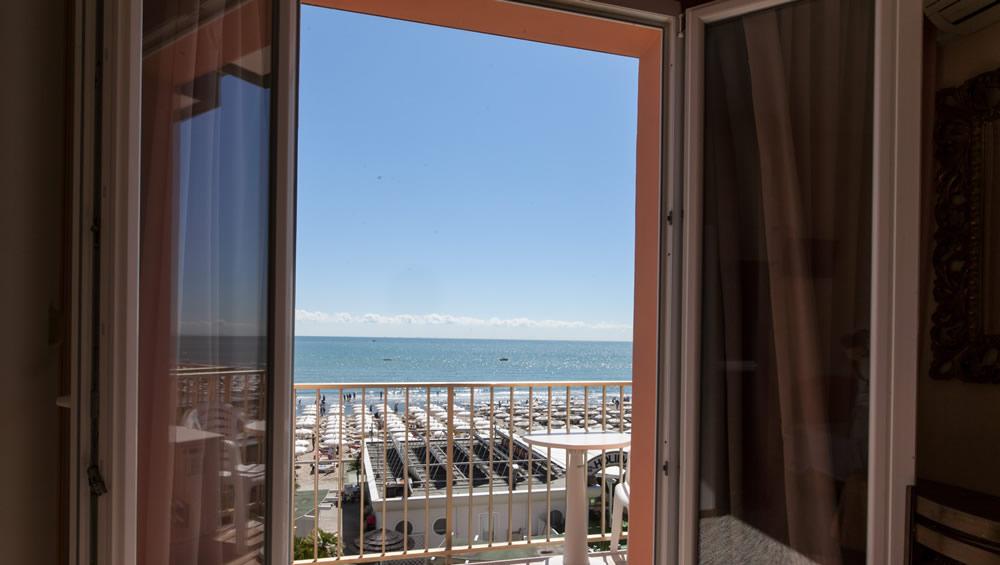 Vista mare da una finestra aperta, con balcone e sedie bianche.