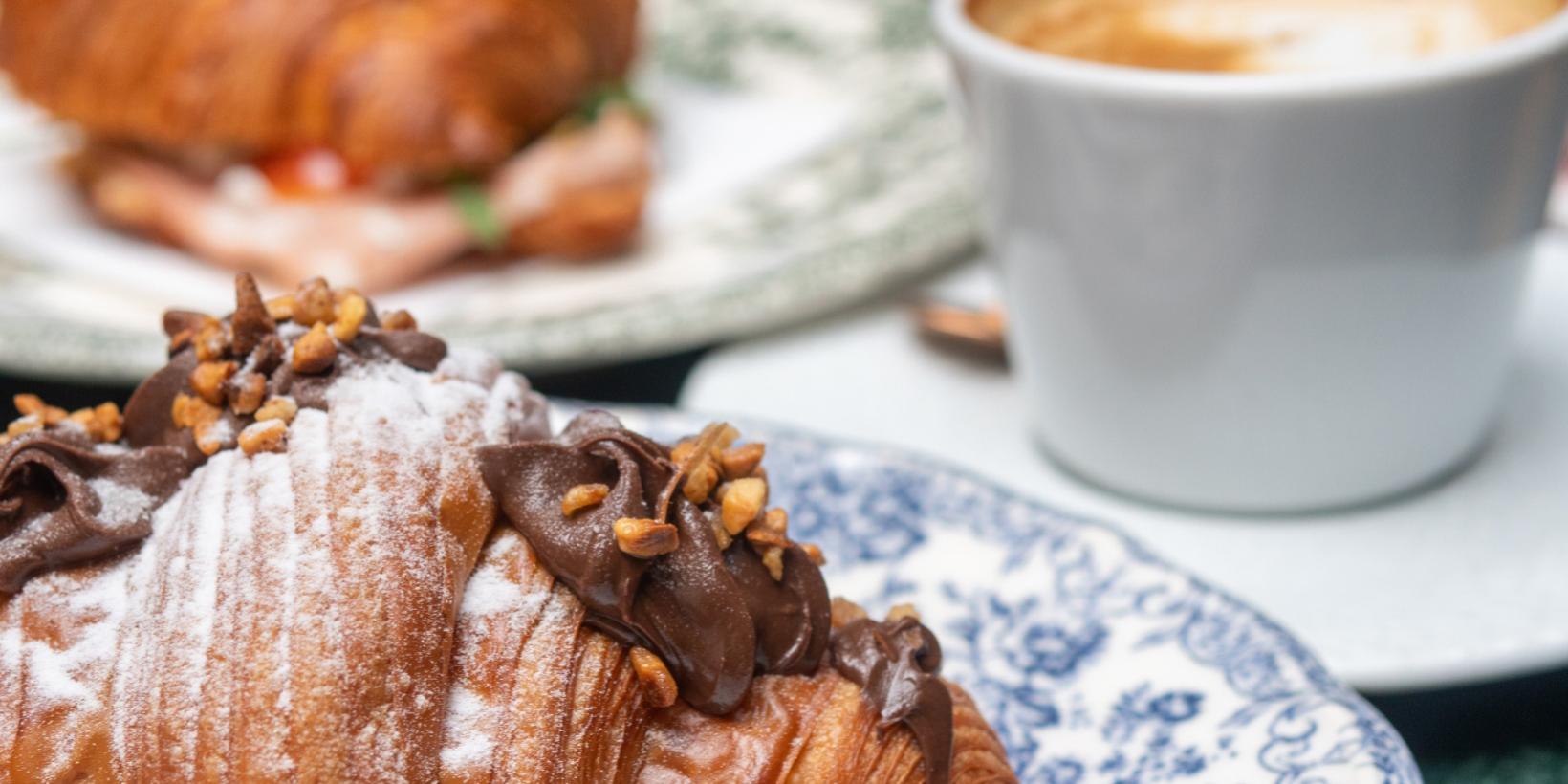Croissant al cioccolato con granella e cappuccino su piatto decorato.