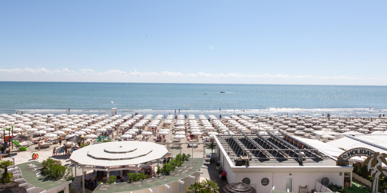 Spiaggia affollata con ombrelloni bianchi e mare calmo.