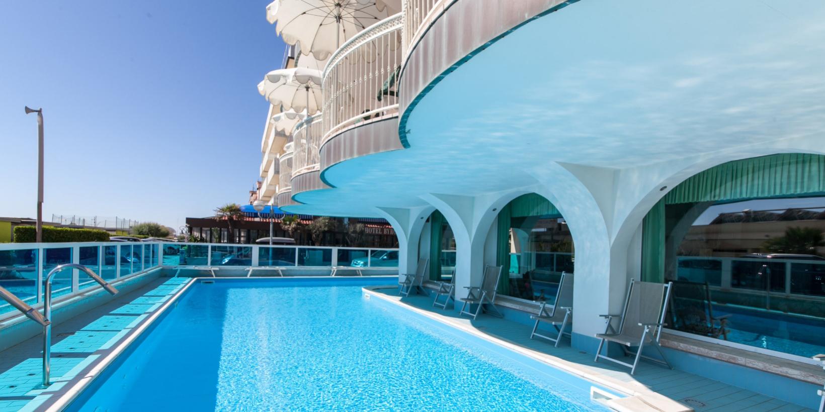 Piscina all'aperto con sedie a sdraio e ombrelloni, accanto a un edificio moderno.