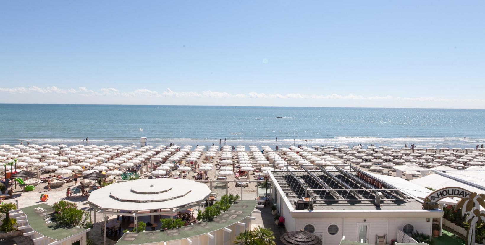 Spiaggia affollata con ombrelloni bianchi e mare calmo sotto un cielo sereno.