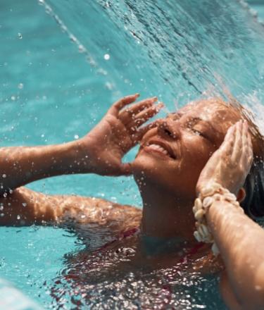Donna felice sotto una cascata in piscina, godendo il sole e l'acqua fresca.