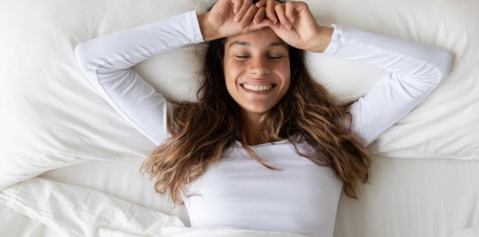 Donna sorridente si sveglia felice in un letto bianco, indossando una maglia bianca.
